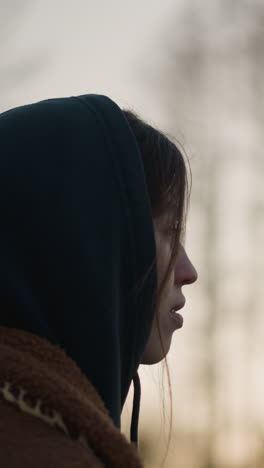 una chica melancólica camina por un parque al atardecer, con un abrigo marrón y una sudadera con capucha, ocultando parcialmente su cara. fondo borroso de árboles y luz solar desvanecida
