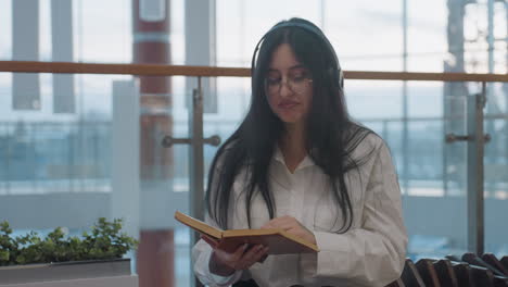 young woman with long black hair, wearing glasses and headphones, seated indoors smiling gently while reading book, subtly swaying with joy, sunlight streaming through glass panels in background