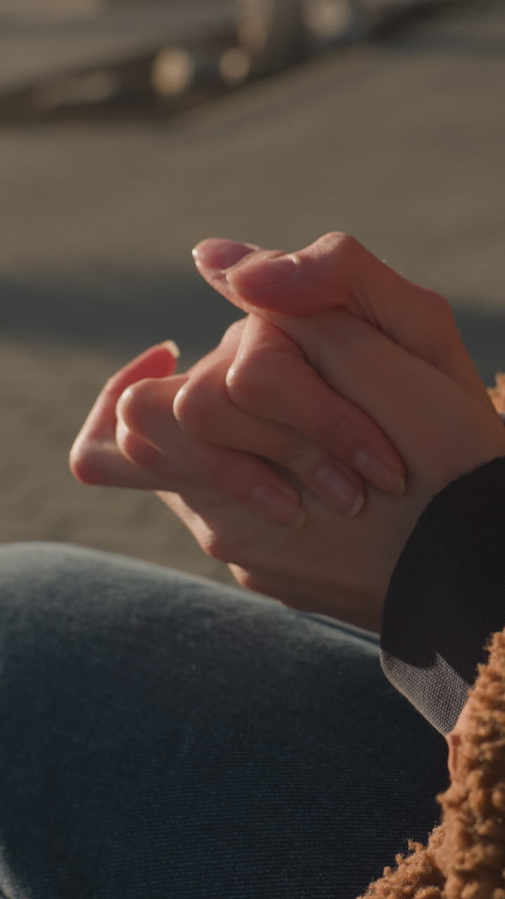 primer plano de una persona sentada con la cara no visible, con pantalones vaqueros azules y un abrigo marrón. las manos están fuertemente juntas en un gesto de ansiedad o pensamiento profundo, transmitiendo tensión o preocupación