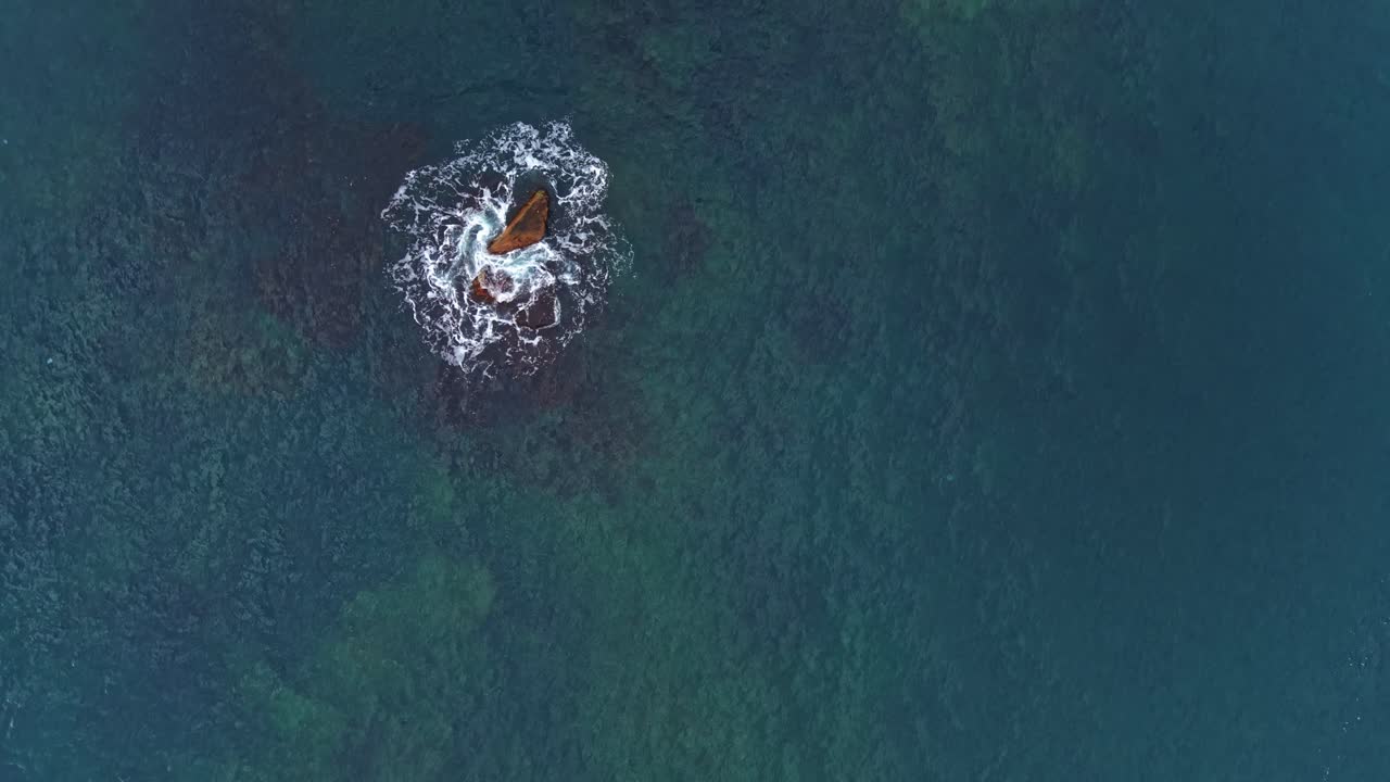 Aerial view of waves crashing around rocky outcrop in clear water