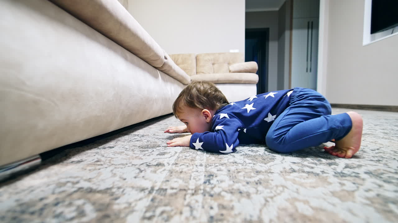 Funny kid falls on the floor to look under the sofa. Lovely toddler trying to get the toy from under the furniture.