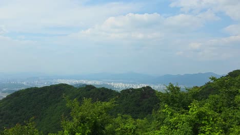 a serene timelapse from namhansanseong captures clouds floating above layers of green, forested mountains, with the sprawling metropolis of seoul visible in the hazy distance.