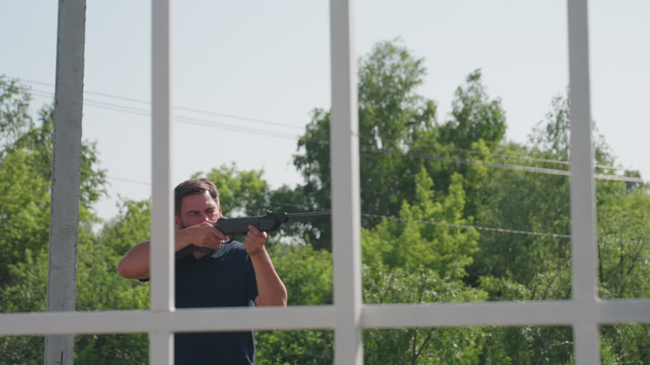 man aiming rifle and shooting pest near farm frame, rural greenery background, sunlight on face, defensive action to protect crops from birds or rodents