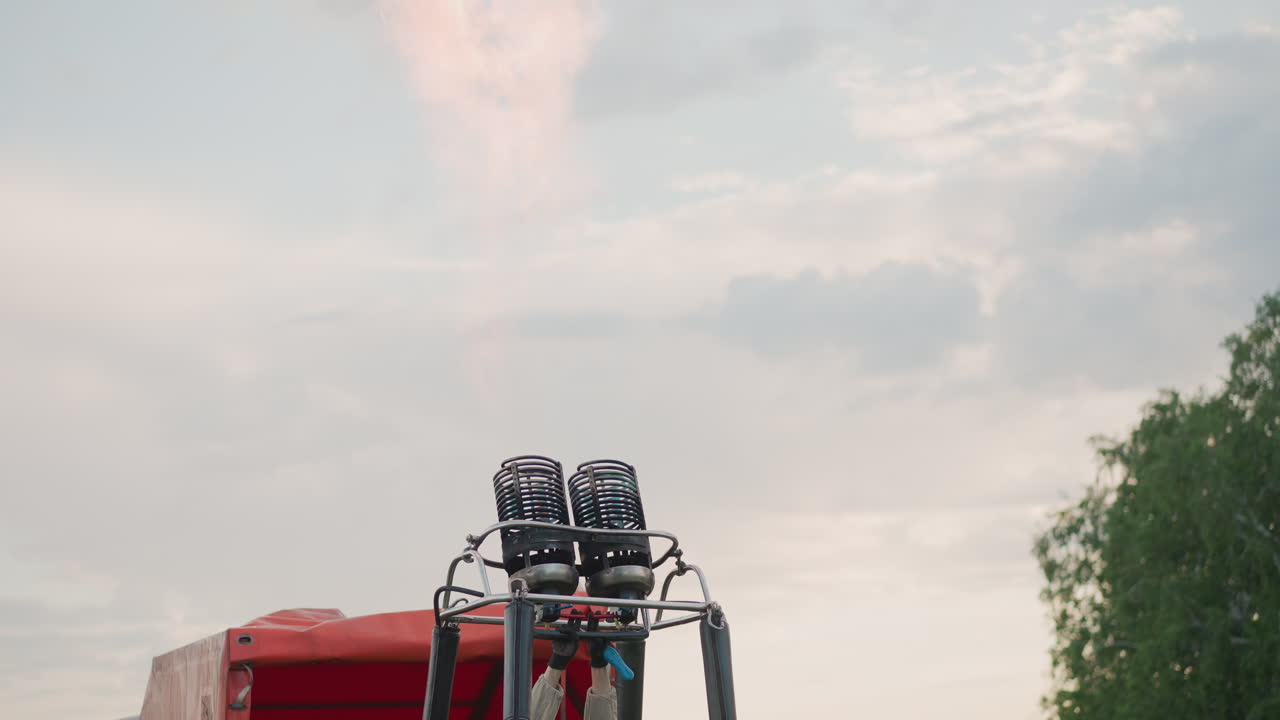 woman igniting metal burner assembly on hot air balloon frame forcing flame higher inside dual burners against red trailer and cloudy sky backdrop over open field at dusk