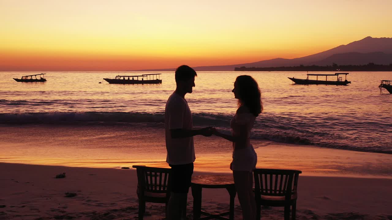 Young couple talking to each other in front of magical lagoon with orange colors of twilight reflecting on calm sea surface filled by Indonesian boats