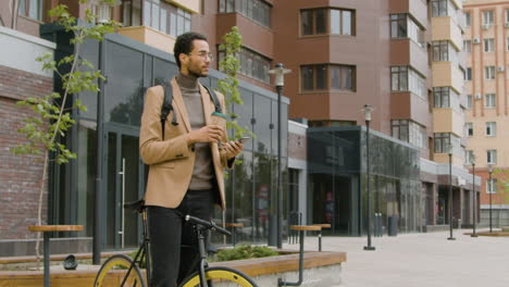 young american man in formal clothes drinking coffee and using mobile phone while standing with his bike in the street