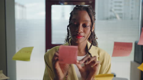 A-young-serious-black-business-woman-in-a-suit-creates-a-board-of-ideas-on-a-glass-wall-gluing-sticks-and-signing-them.