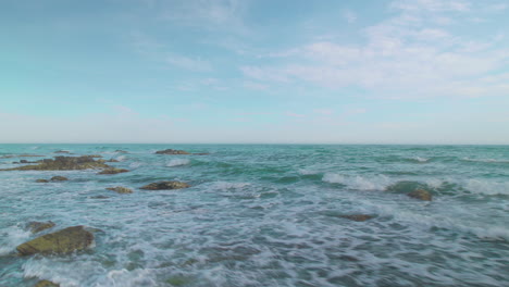 aérea moviéndose diagonalmente en una playa rocosa, con nubes dispersas y olas suaves en el fondo