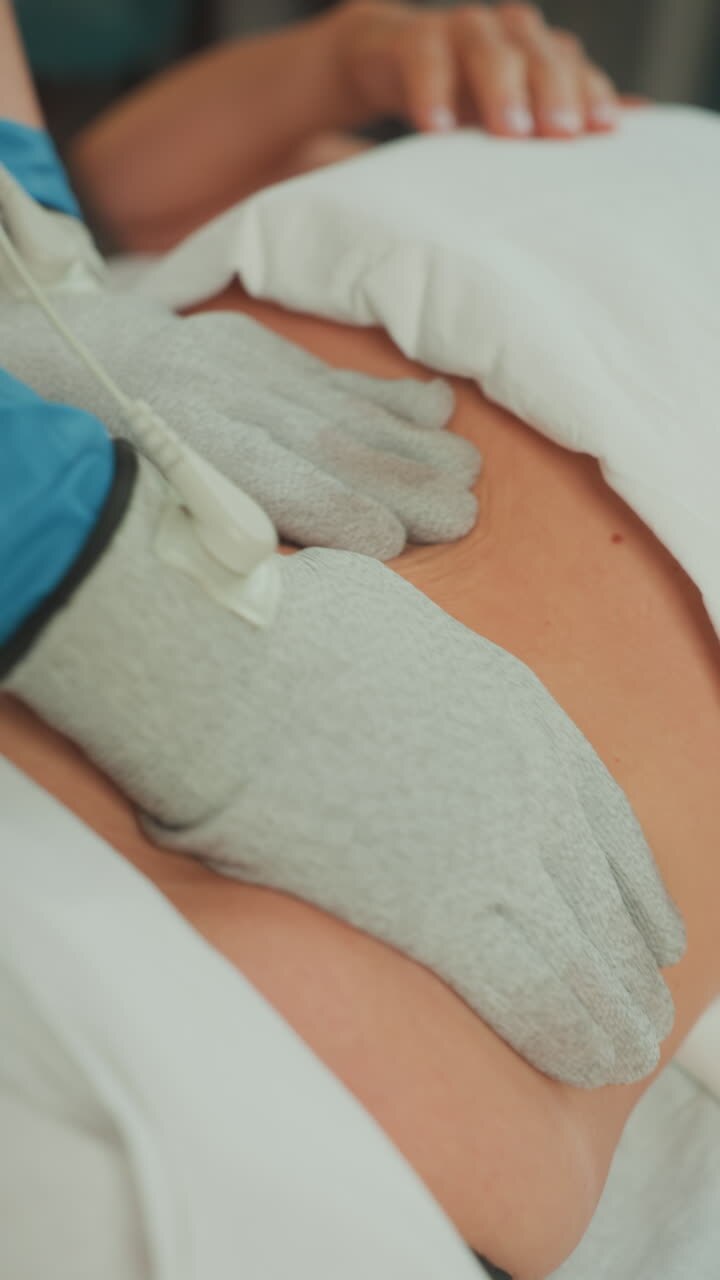 donna caucasica addome massaggio guanti microcorrente, estetista che indossa guanti conduttivi esegue un massaggio terapeutico a microcorrente sulla parte inferiore dell'addome, lenzuola bianche cliniche, elettrodo cablato