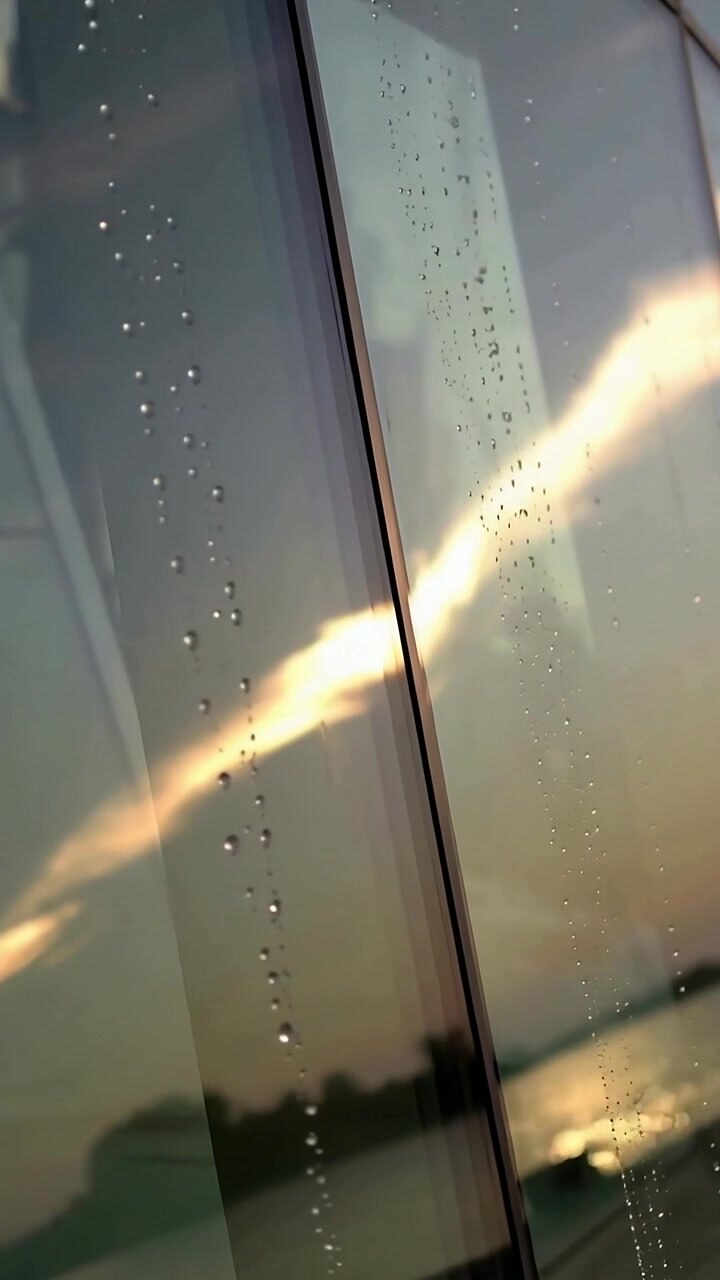 Vertical video: Shifting glass panel with seam and drops showing sun streak over bay, camera moving