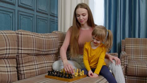 Feliz-Joven-Madre-Enseñando-A-Su-Hija-Pequeña-A-Jugar-Ajedrez-En-Una-Tabla-De-Madera-En-El-Sofá-De-Casa