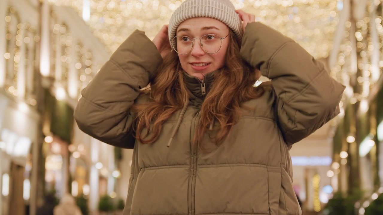 Full shot white woman holding backpack and plush toy, adjusting beanie then smiling, playful shopper vibe in holiday market, warm lights and decorated storefronts, confident stride in puffer coat