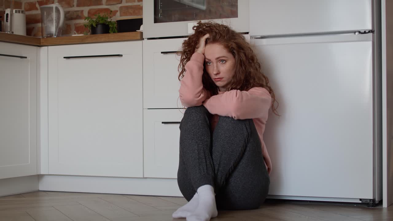 Sad young caucasian woman sitting sad on floor in the kitchen.