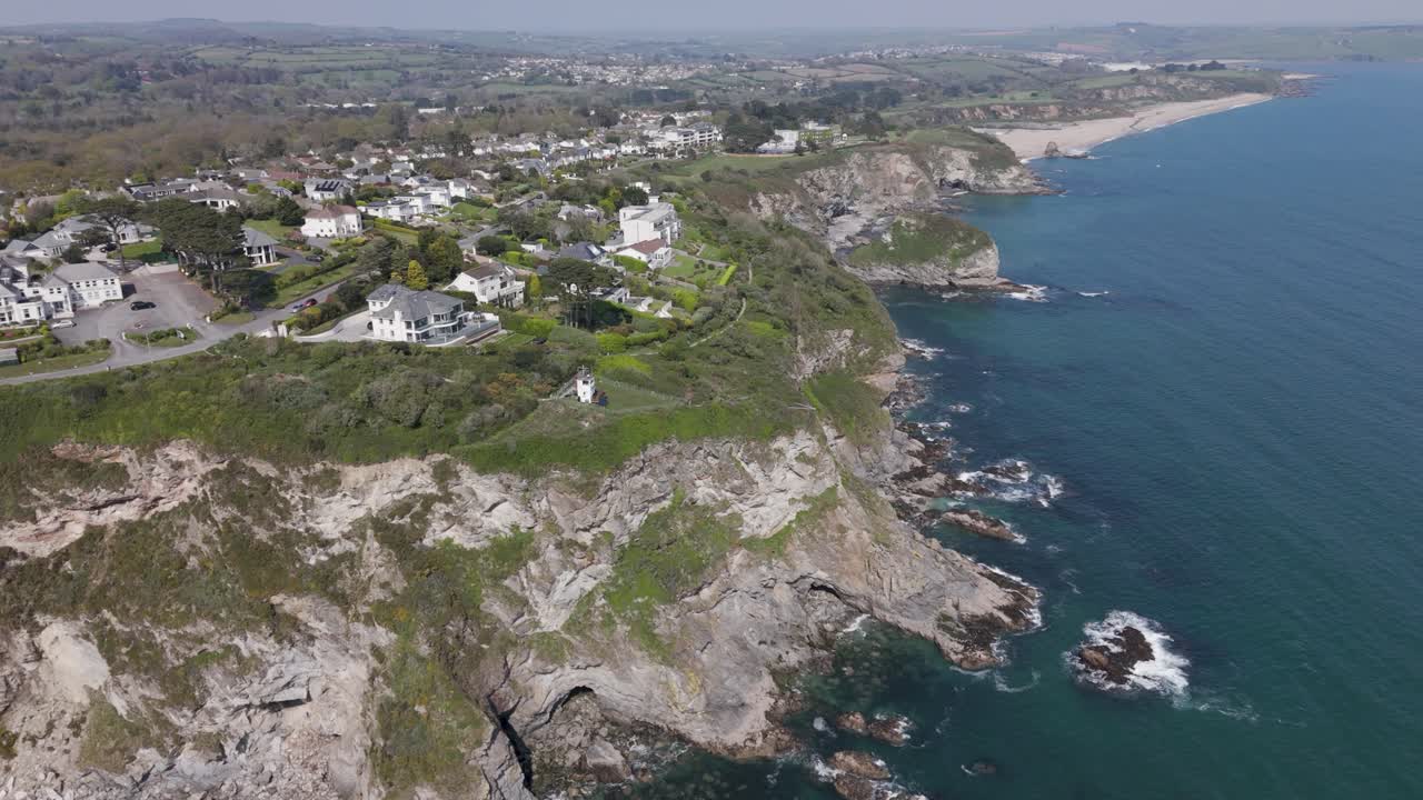 Aerial push-in along rugged Cornish cliffline where modern luxury holiday villas with lawns and footpaths overlook turquoise Atlantic waves and dramatic rocks