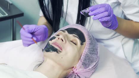 beautician is holding brush near woman's face and applying black facial mask in beauty cabinet. attractive female in medical cap relaxing with closed eyes and enjoying spa procedures.