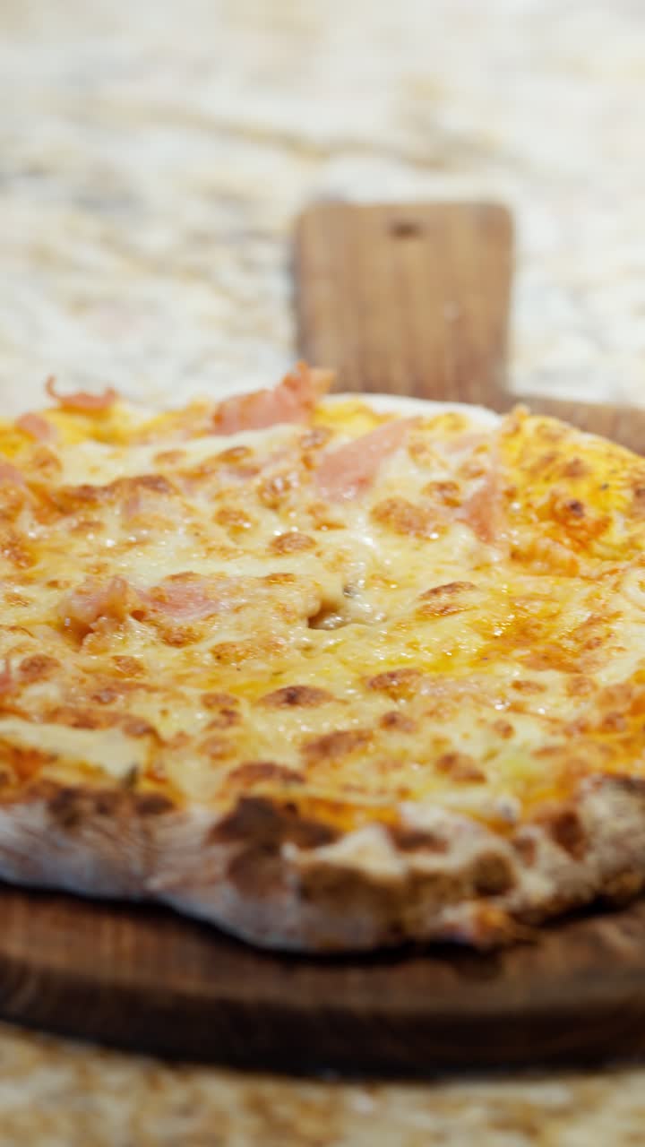 Vertical close-up pan shot reveals a delicious hot freshly baked Italian pizza with melted cheese and ham, served on a rustic wooden board in a restaurant