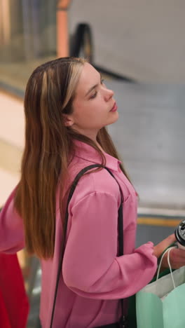 mujer con vestido rosa sosteniendo taza de café y bolsas de compras mientras está de pie en una escalera mecánica mientras desciende en un centro comercial bien iluminado, parece cansada, con diseños de paredes coloridas y barandillas de vidrio visibles