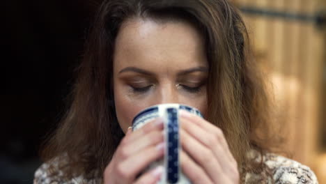 frontal close up fhd shot of a woman holding a cup with both hands and drinking freshly poured hot tea, savoring the taste with a wooden house in the background