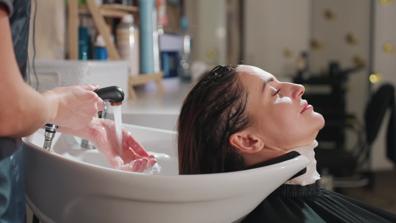 Kunde under hårvask, professionel stylist justerer vand og vask for kundens komfort, kunden slapper af i ergonomisk designet salonvask, mens stylisten kontrollerer vand- og vaskefunktioner.