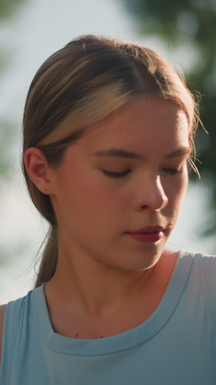 Young lady in light blue top adjusts posture while focusing downward with hair tied back, with background features blurred view of green leaves
