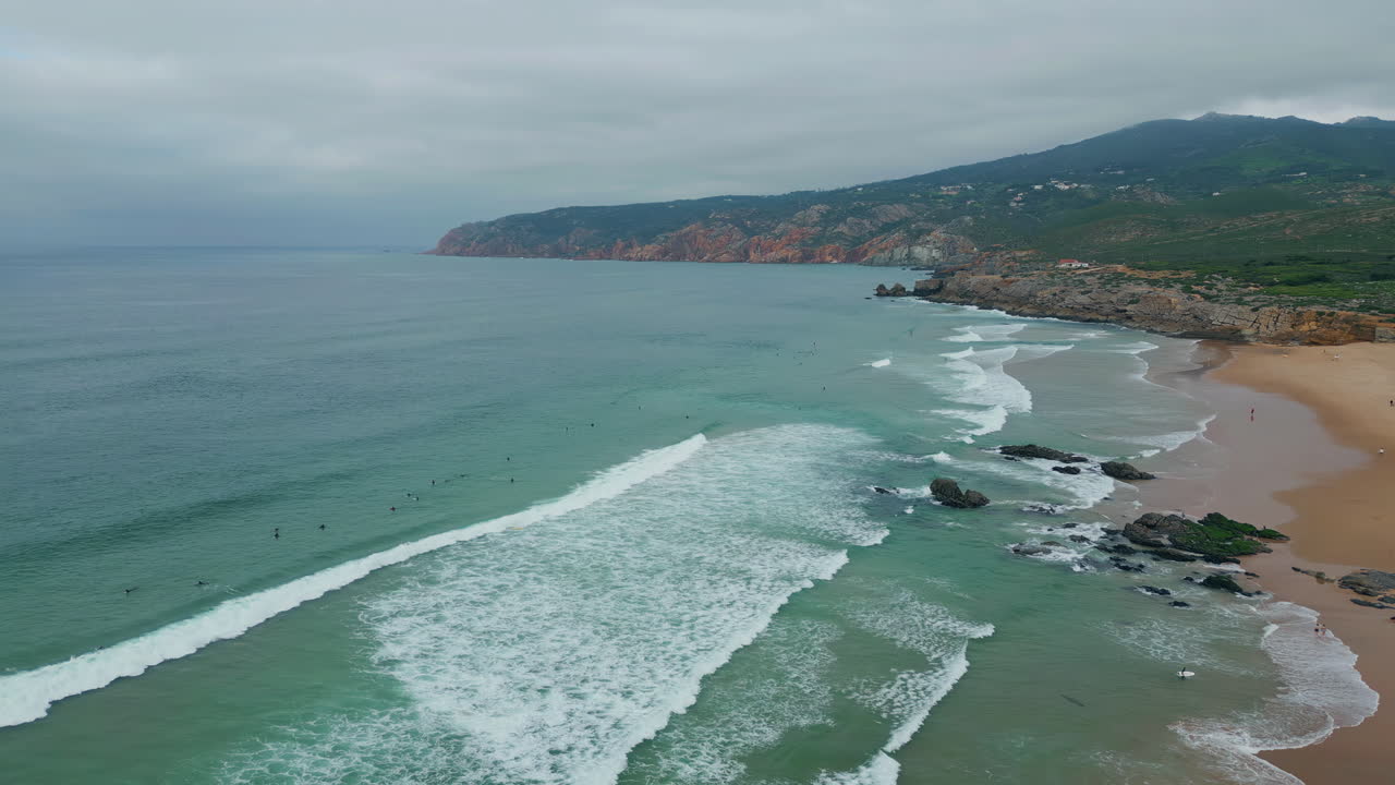 Vista aerea di onde oceaniche che si infrangono sulla riva in una giornata nuvolosa. Scogliere frastagliate sopra i surfisti.