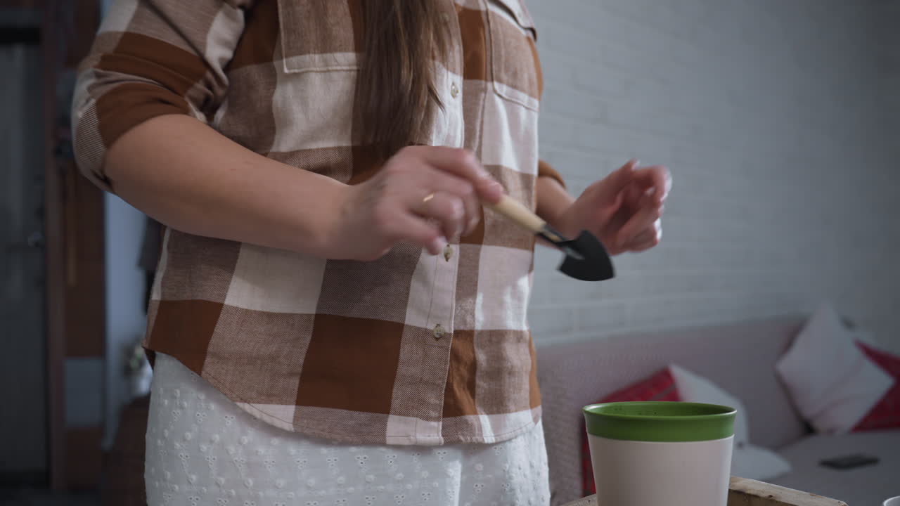 Plant loving creative twirling joyfully with small shovel while working soil into planter on glass table amid lush indoor plants in bright white room radiating playful gardening energy