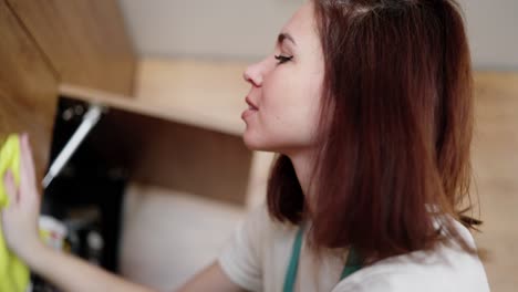 retrato en primer plano de una confiada mujer de limpieza morena con un delantal blanco y una camiseta blanca que limpia los gabinetes superiores en una cocina de color madera con un trapo amarillo durante la limpieza