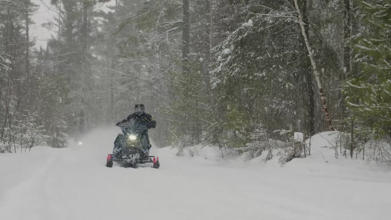 Ormanın arkasından çıkan iki kar motosikleti, savrulan karların içinden hızla geçiyor.