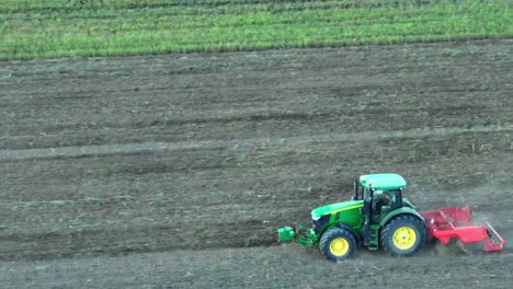 green colored tractor with yellow tires plowing on brown field in nature - aerial view