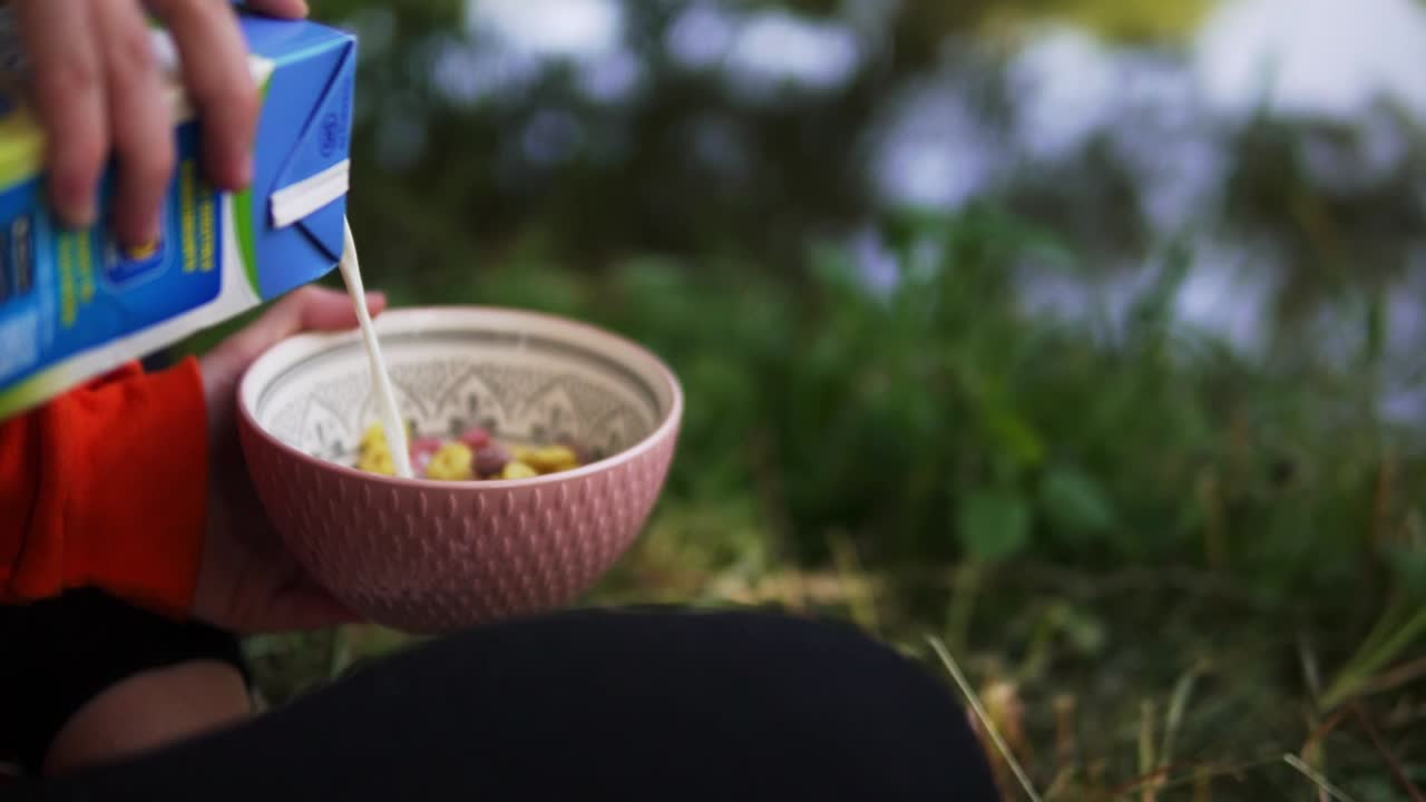 강이나 호수 앞 풀밭에 앉아 야외 캠핑에서 시리얼이 든 그릇에 우유를 붓는다