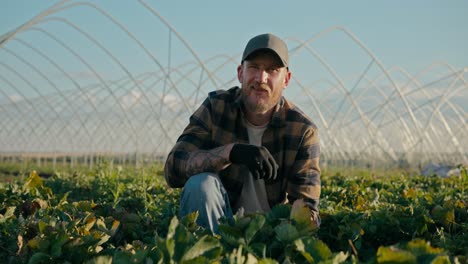 Portrait-of-a-confident-farmer-guy-in-a-cap-and-plaid-shirt-posing-while-sitting-among-growing-crops-on-a-farm