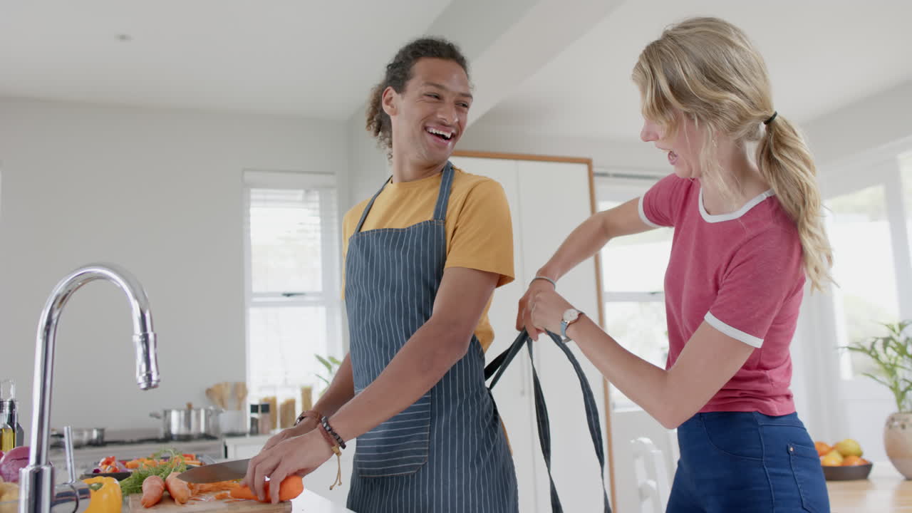 Happy diverse couple preparing fresh vegetables and wearing aprons in kitchen, slow motion