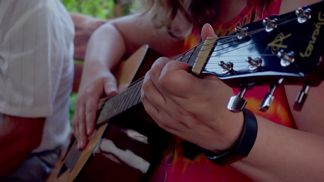 Ripresa ravvicinata in movimento di una donna che suona la chitarra acustica durante una festa estiva all'aperto con la famiglia a Daugavpils, Lettonia, catturando un'atmosfera musicale pacifica.