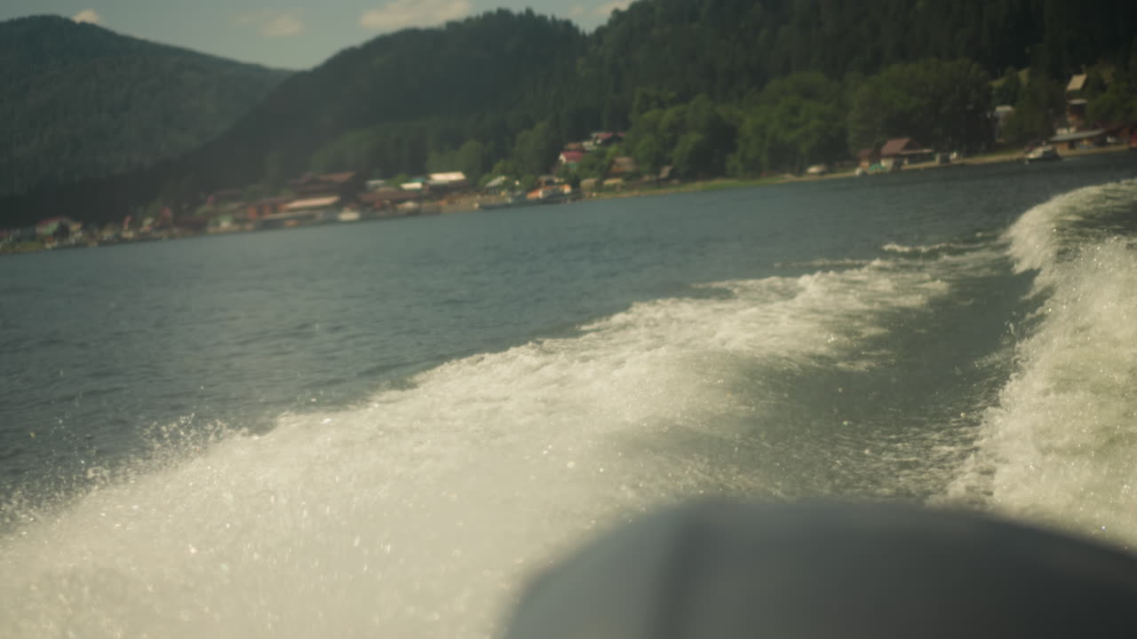 всплески водных волн за быстрым кораблем на морской воде против деревни на побережье вблизи. пенообразная тропа на поверхности лазурного озера после плавания на моторной лодке