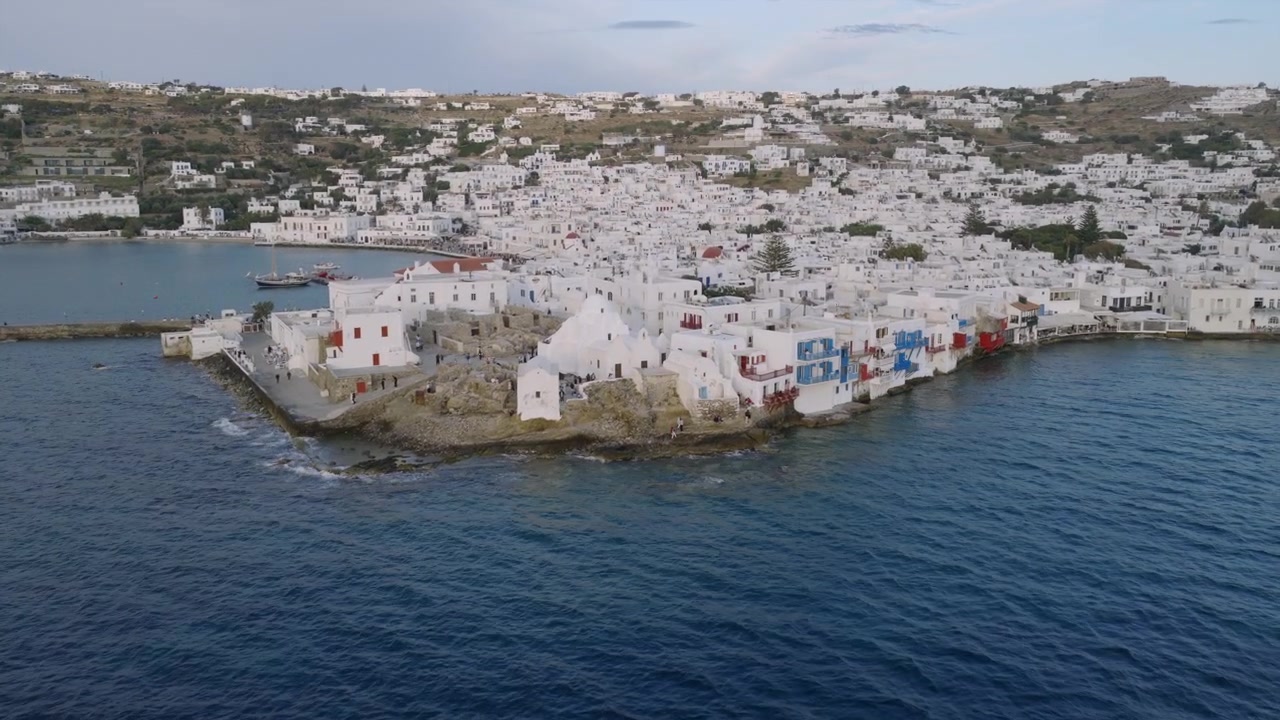 Ampia vista aerea della città di Mykonos dall'alto del profondo blu dell'Egeo, con un traghetto vicino al molo ed edifici bianchi che ricoprono le colline oltre.