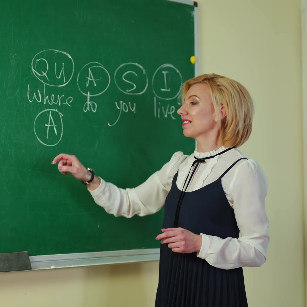 Teacher near the blackboard. Woman writes with white chalk on a green chalkboard at school. Female teacher explains the rule in the classroom.