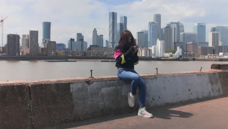 Full-shot-of-a-girl-sitting-while-chatting-on-the-phone-near-the-river-with-a-city-in-the-background
