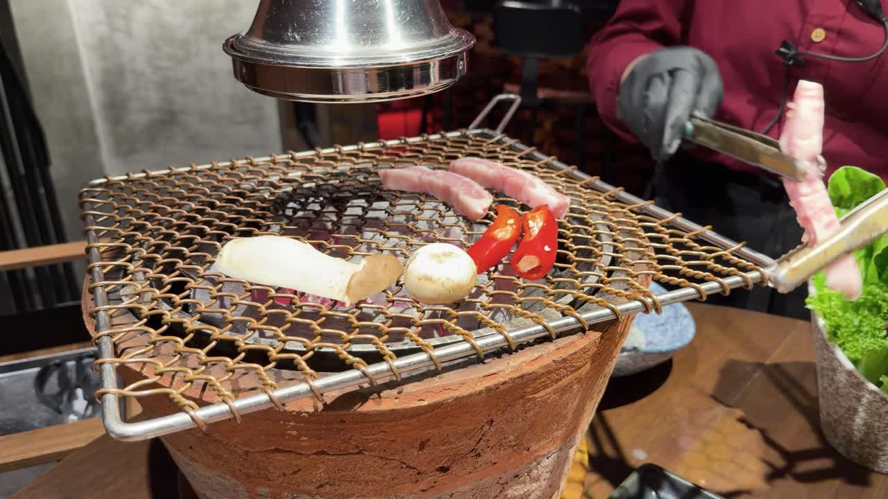 Gloved chef grills pork, mushrooms, and peppers on tabletop charcoal grill under bright indoor lighting