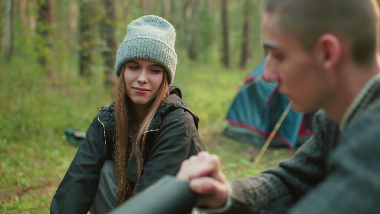 Close up of man pumping air into object while woman stands nearby smiling warmly, both engaged in camping activity with tent and forest visible in background