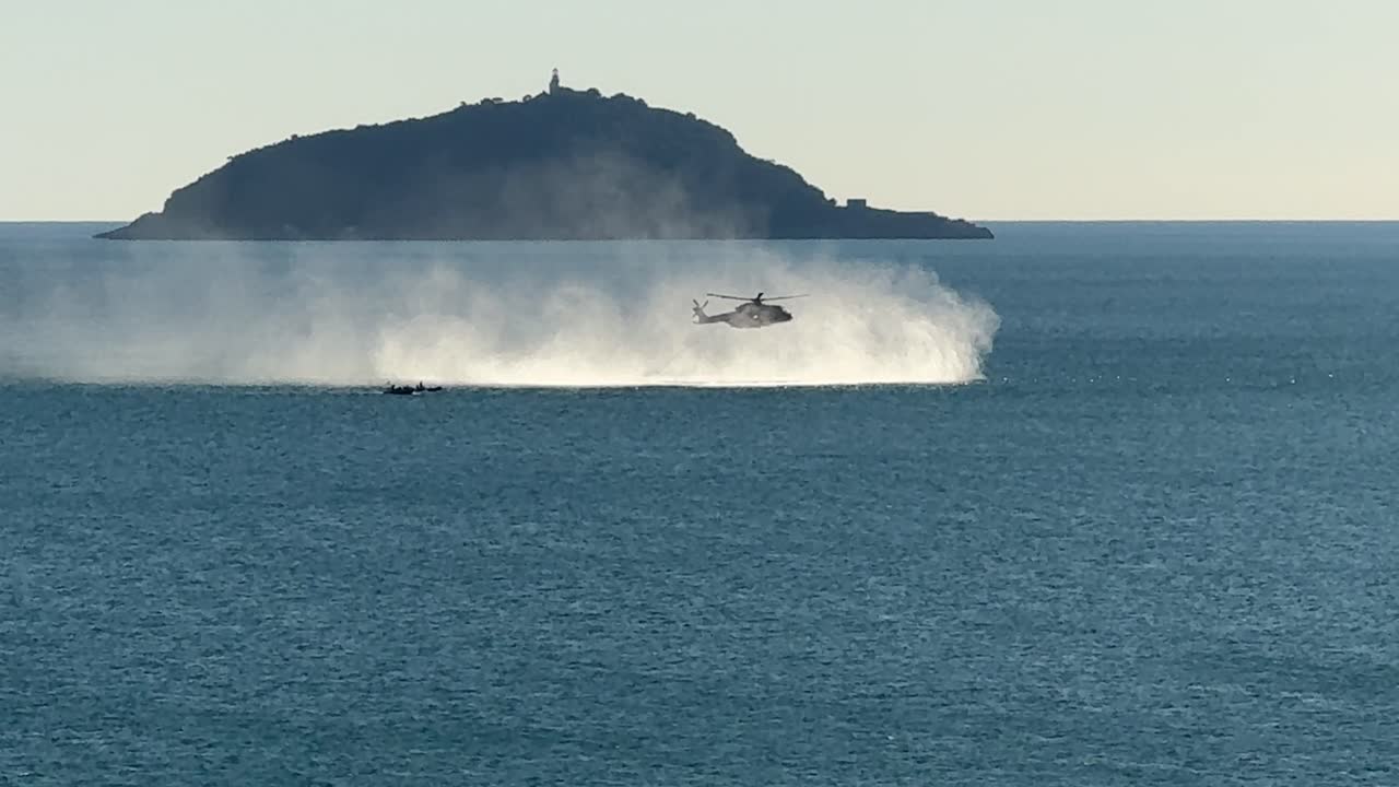 Italian Navy helicopter conducts maritime training exercise over water near Tino Island in the Gulf of La Spezia, creating dramatic spray under clear Mediterranean skies