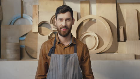 portrait of caucasian bearded man in apron looking at camera and smiling in carpentry workshop