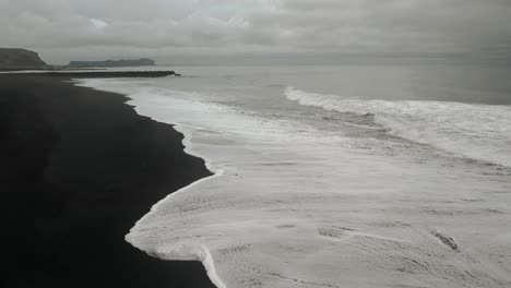 Antena-Cinematográfica-Playa-De-Arena-Negra-Olas-Del-Océano-Rompiendo-En-La-Orilla,-Paisaje-Oscuro-Y-Cambiante,-Vik,-Islandia