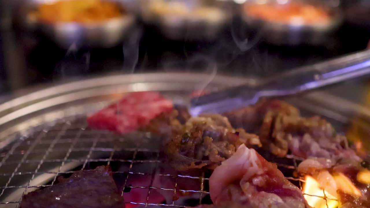 Irisan tipis daging sapi mendesis di atas panggangan arang panas dengan asap yang mengepul dan penjepit.