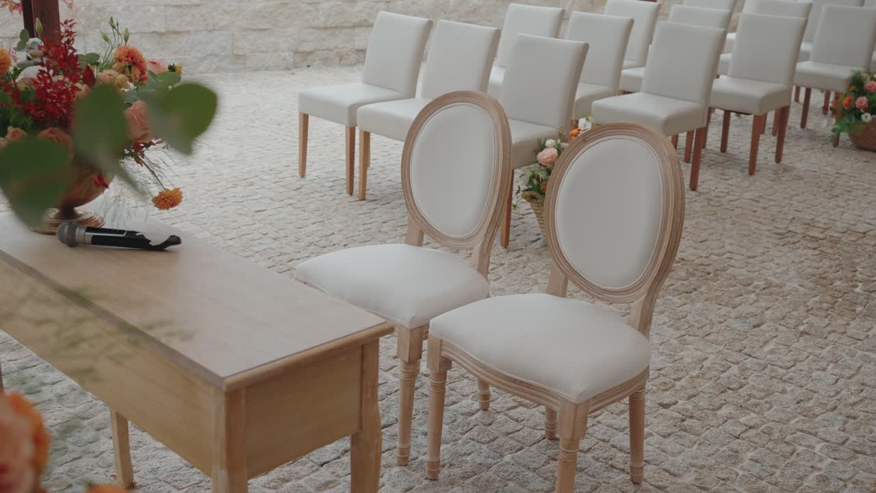Light wooden table with black handheld microphone placed on top beside two oval back upholstered chairs on cobblestone floor, with rows of white chairs and floral baskets in background