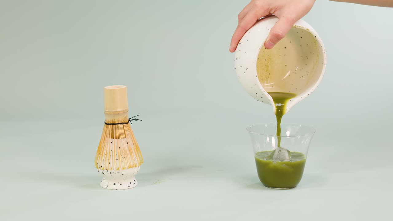 Hand pours matcha tea from ceramic bowl into iced glass, soft lighting, minimalist studio setting