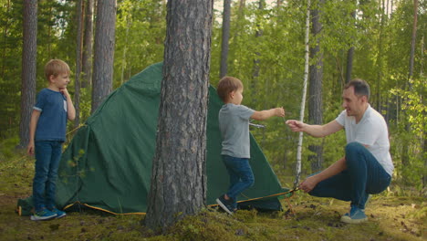 the children together with their father set up a tent for the night and camping in the forest during the journey. a man and two children 3-5 years old together in a hike gather a tent in slow motion