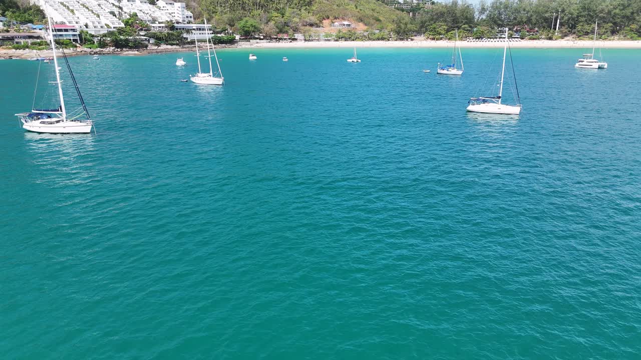 Drone footage captures serene boats anchored in Phuket's turquoise waters, showcasing a tranquil coastal scene with lush surroundings