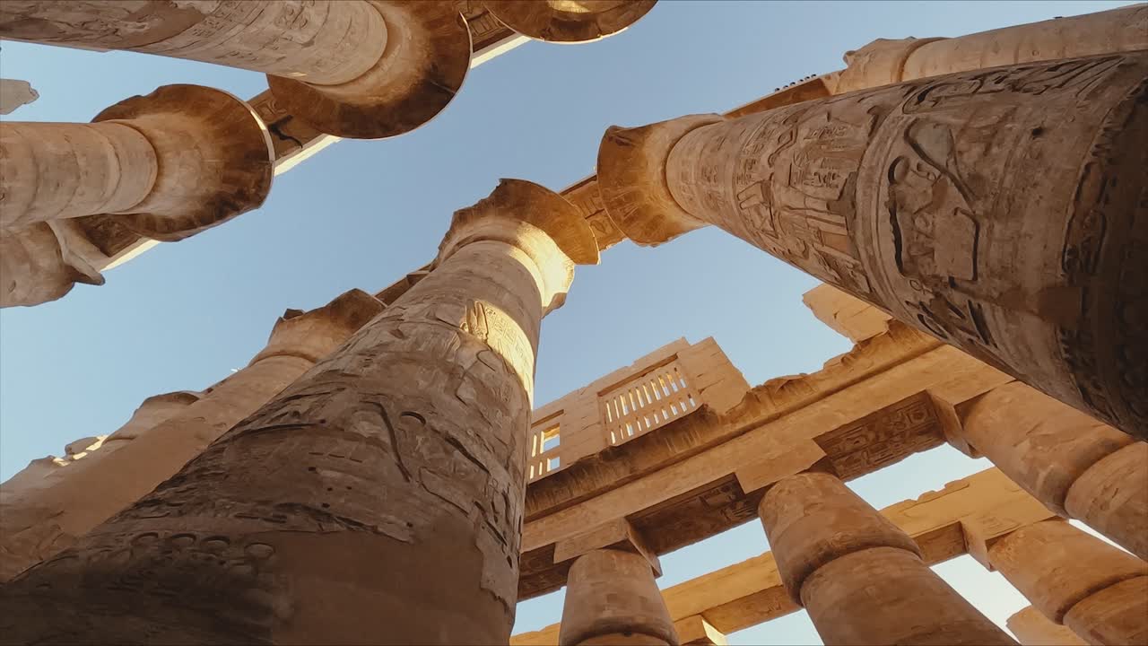 Low-angle view of towering stone columns at Karnak Temple in Luxor, Egypt. The intricate hieroglyphics and monumental architecture are bathed in warm sunlight.