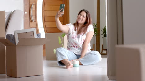 Feliz-Mujer-Birracial-De-Talla-Grande-Tomándose-Selfie-Sentada-En-El-Suelo-De-Una-Nueva-Casa,-Espacio-Para-Copiar,-Cámara-Lenta