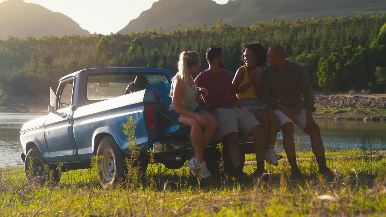 Friends With Backpacks Sitting On Tailgate Of Pick Up Truck On Road Trip By Lake In Countryside